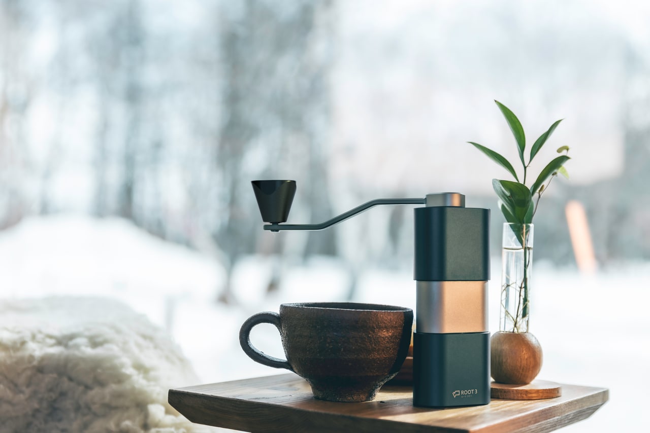 ROOT3 coffee grinder with its manual handle, photographed beside a coffee cup
